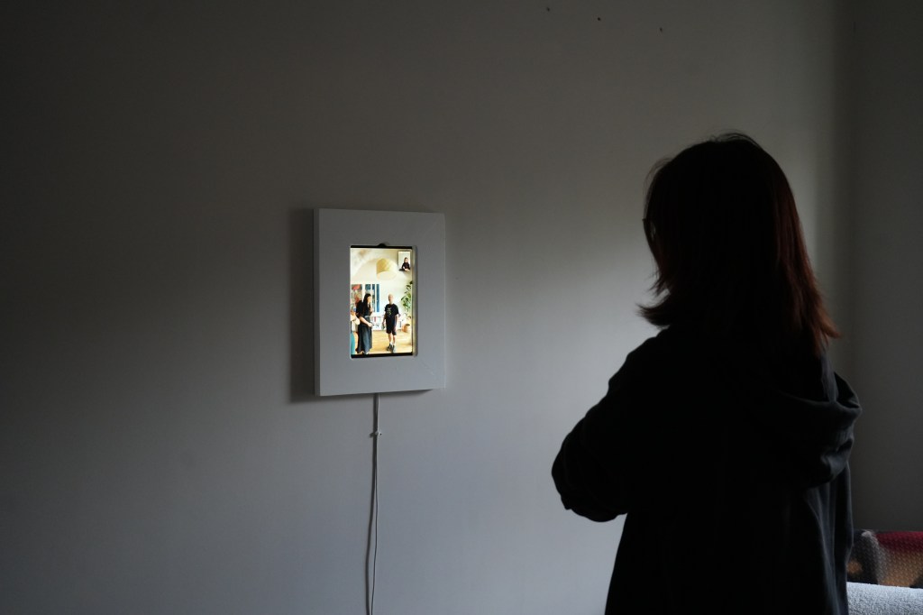 A person stands with their back to the camera, observing a digital photo frame on the wall displaying an image of two people in a bright interior space.