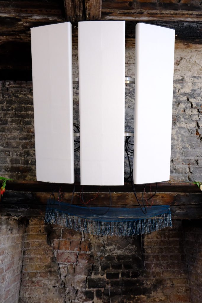 Three tall, white acoustic panels mounted on a wooden beam in an industrial space, with a blue fringe hanging below.
