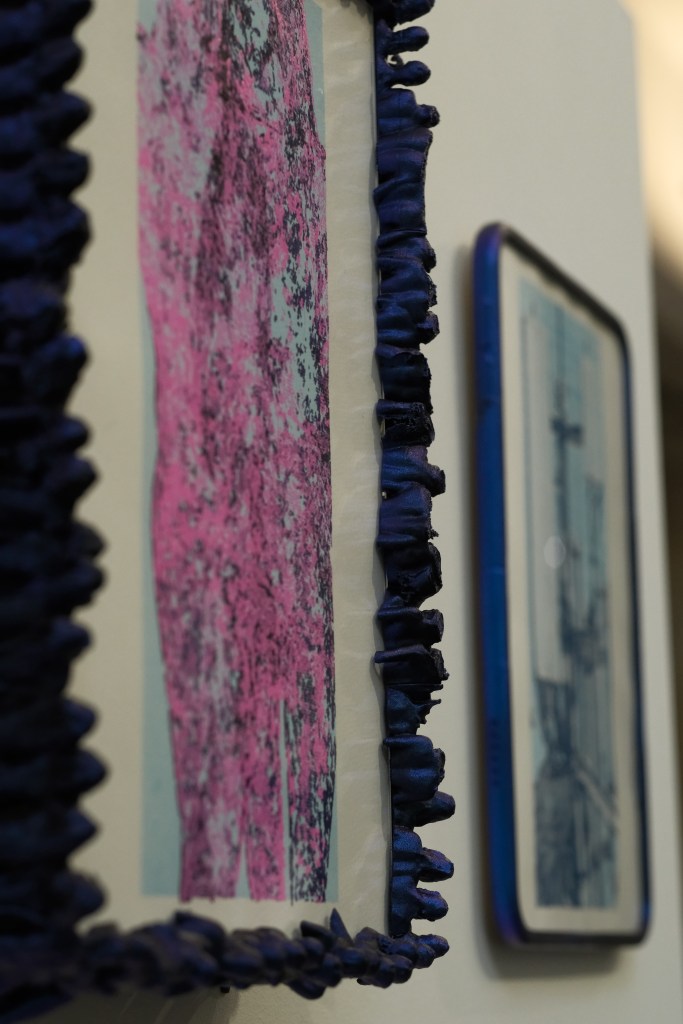 Close-up view of a colorful artwork, featuring a pink and teal abstract design framed with a textured blue border, alongside a second framed piece in the background.