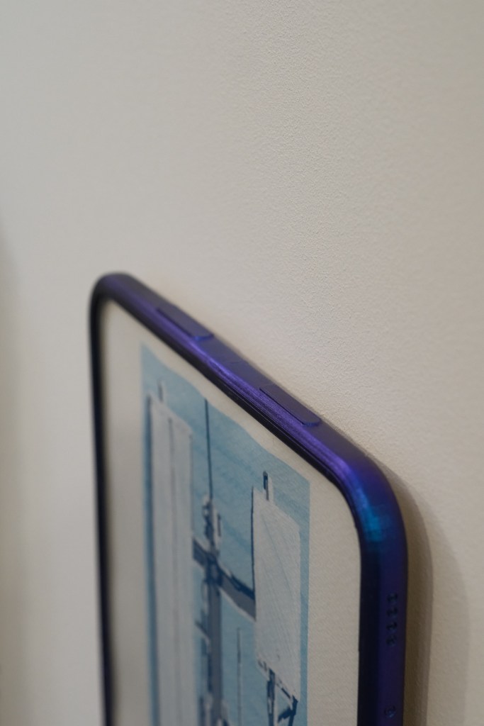 Close-up of a smartphone with a purple case, positioned against a plain white wall. Part of a blue-colored artwork is visible on the screen.