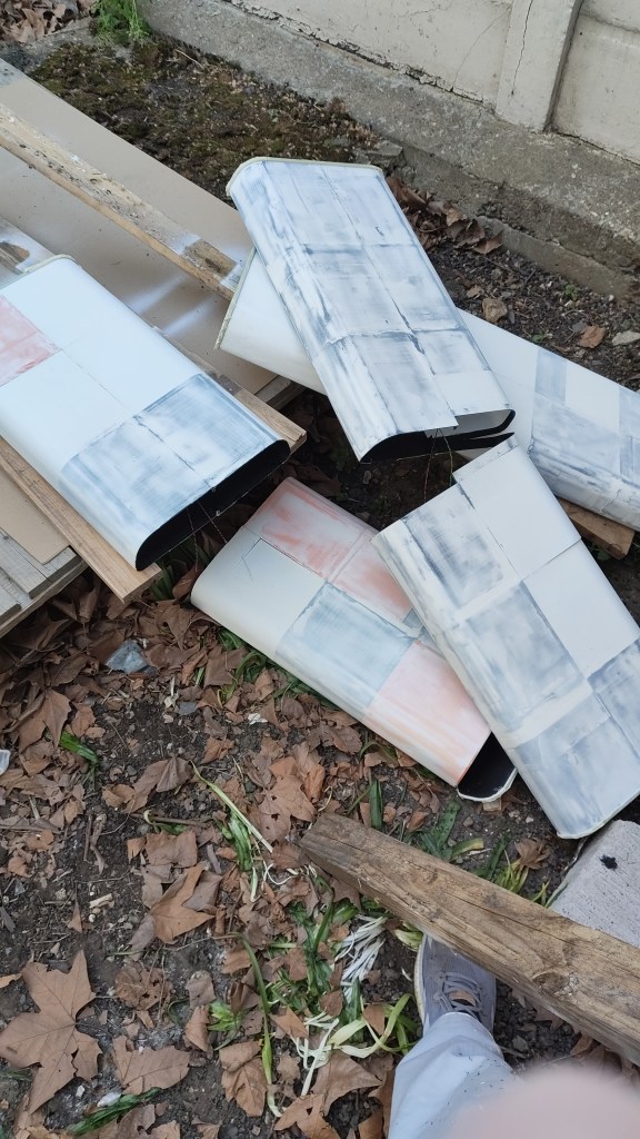 A pile of partially painted metal sheets surrounded by fallen leaves and plant growth on the ground.