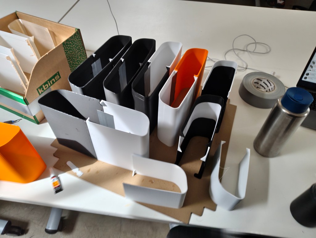 An assortment of colorful and monochrome paper containers on a workspace, including black, white, and an orange container, alongside crafting materials and a metal water bottle.