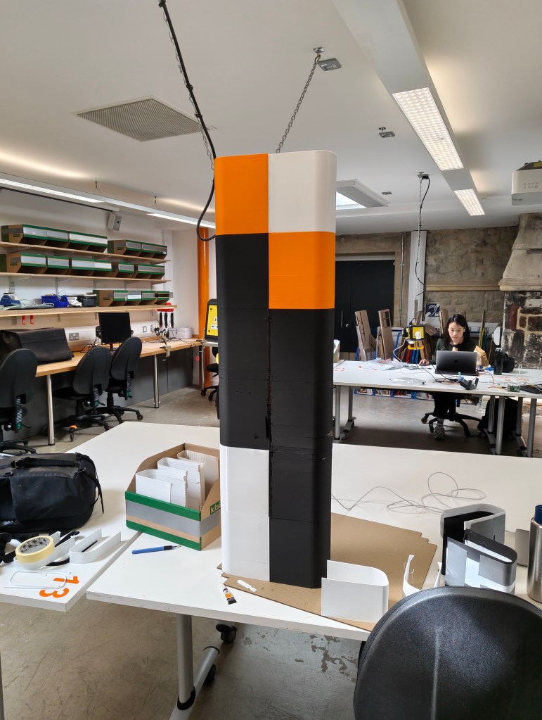 A tall, multi-colored structure featuring orange, black, and white segments, set against a workspace with desks, computers, and various materials. A woman is seen working on a computer in the background.