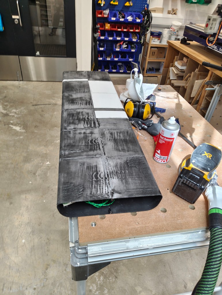 A workshop scene featuring a long, black wooden board with a partially visible green wire underneath. The board is resting on a workbench with tools, including headphones, spray can, and a power tool nearby, while storage bins are seen in the background.