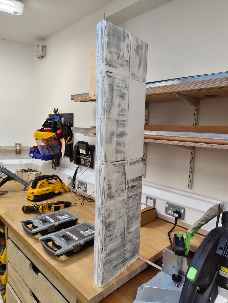 A vertical slab of material with a textured, gray surface, positioned on a workbench with various tools and storage containers in a workshop setting.