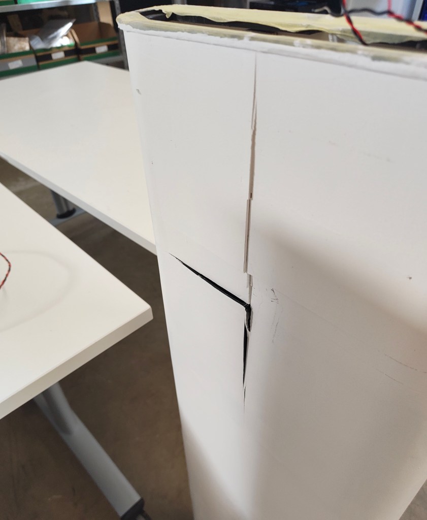 A close-up of a damaged white cardboard box showing a prominent crack on the surface, with a blurred workspace in the background featuring white tables.