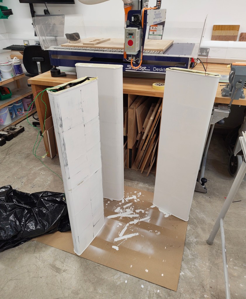 Three vertical panels with white paint, standing on a cardboard base in a workshop, with spilled paint and a black tarp on the floor.