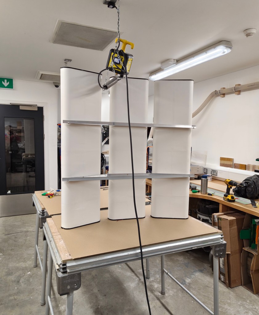 A workspace featuring a white display shelf structure hanging from a ceiling hoist, with tools and equipment visible on the surrounding workbenches.