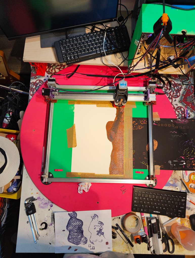 Overhead view of a workspace with a cutting machine, a computer monitor, and various art supplies. The workspace features a colorful patterned table, tools, and a partially completed artwork on the printer.
