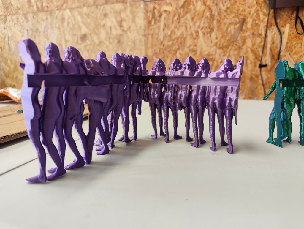 A line of purple 3D-printed human figures, with a few green figures displayed on the right side. The figures are partially obstructed by a horizontal purple band connecting them.