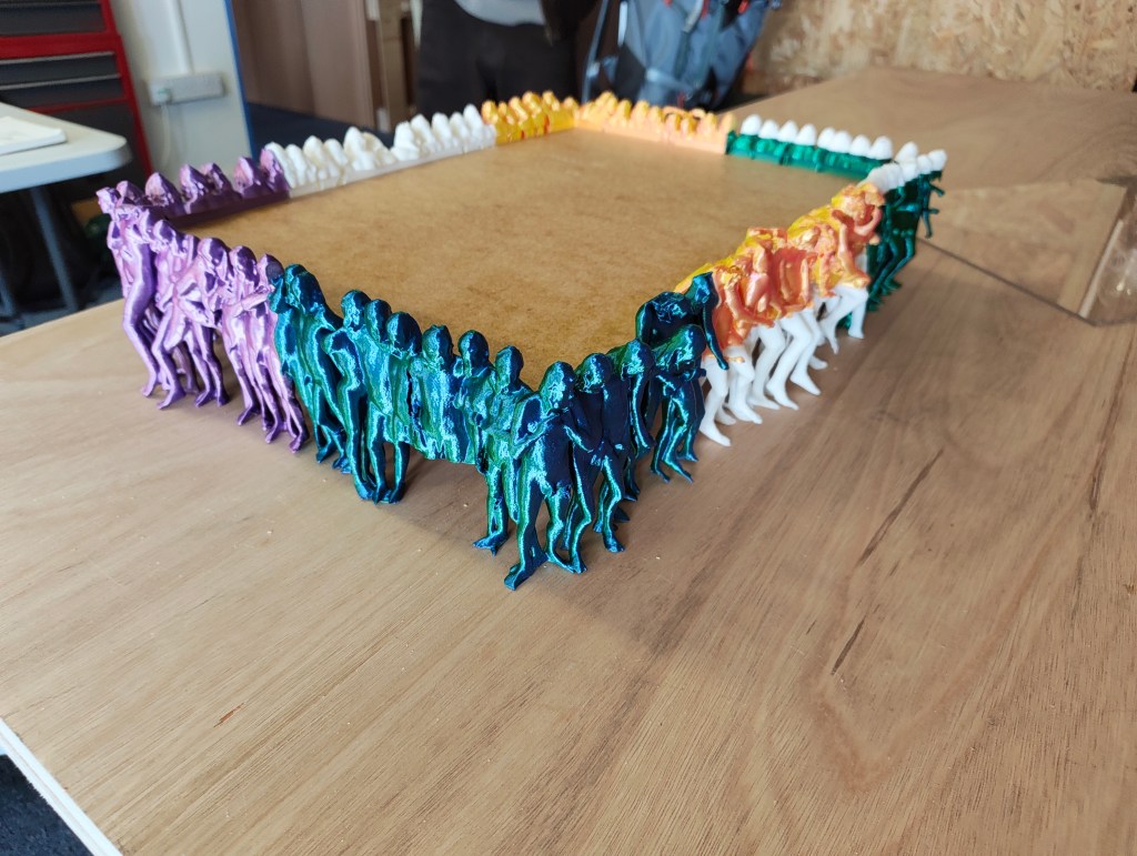 A colorful arrangement of 3D-printed figures, displayed in a square formation around a wooden base, showcasing various colors including purple, green, orange, and white.